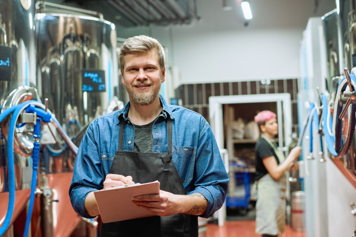 A bearded male worker standing in a brewery while taking notes on a clipboard.