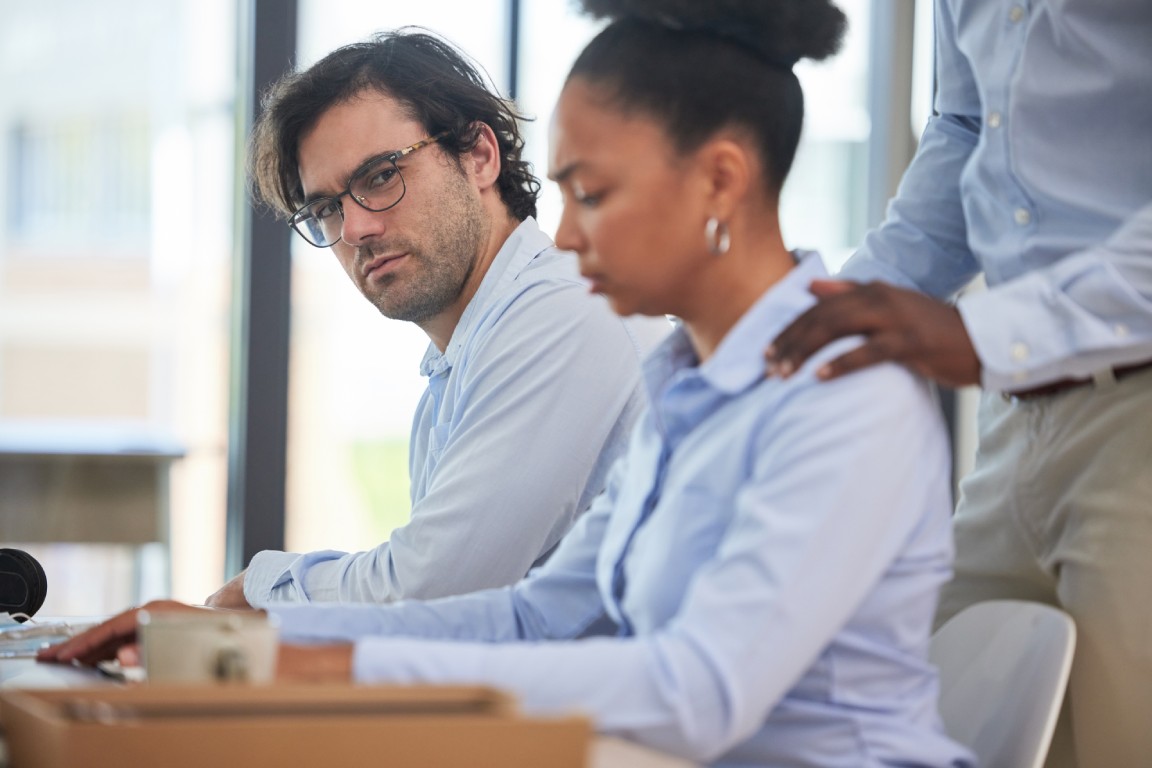 A female employee in an uncomfortable workplace setting with a male coworker and someone touching her shoulders.