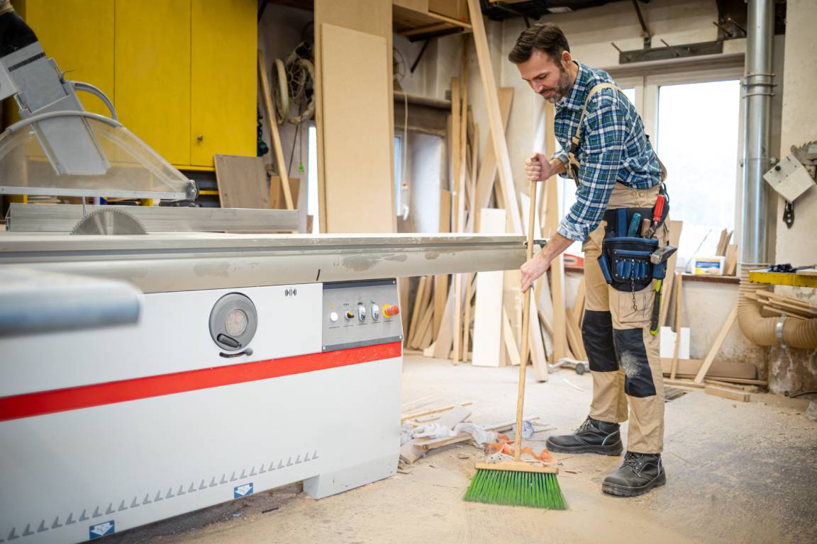 A man wearing coveralls and sweeping wood dust off of his workshop floor with a broom after a project.