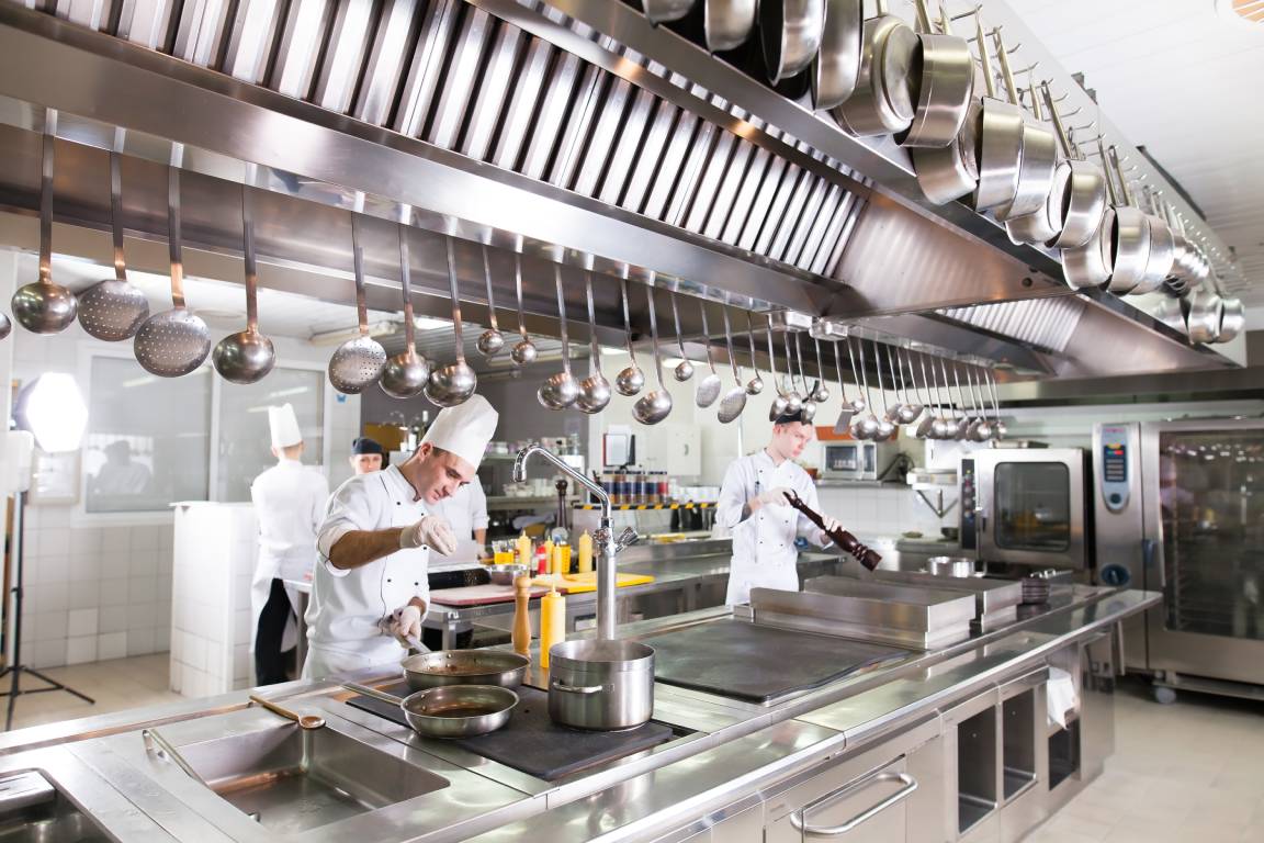 A group of chefs cooking in a large commercial kitchen. The kitchen has stainless steel appliances, counters, and equipment.