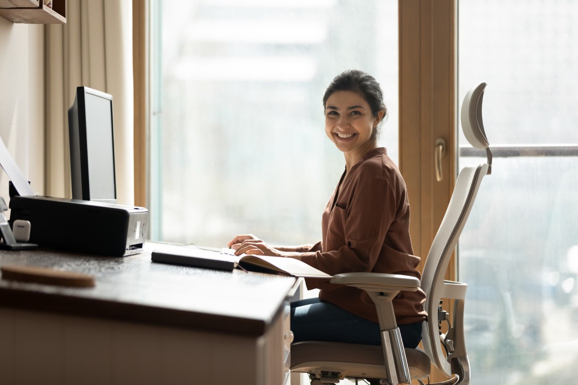 A smiling woman sits in a chair, using a computer monitor inside an office environment with large windows in the background.