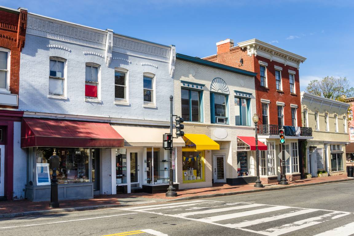 A street with a crosswalk alongside a brick sidewalk lined with several traditionally designed commercial buildings.