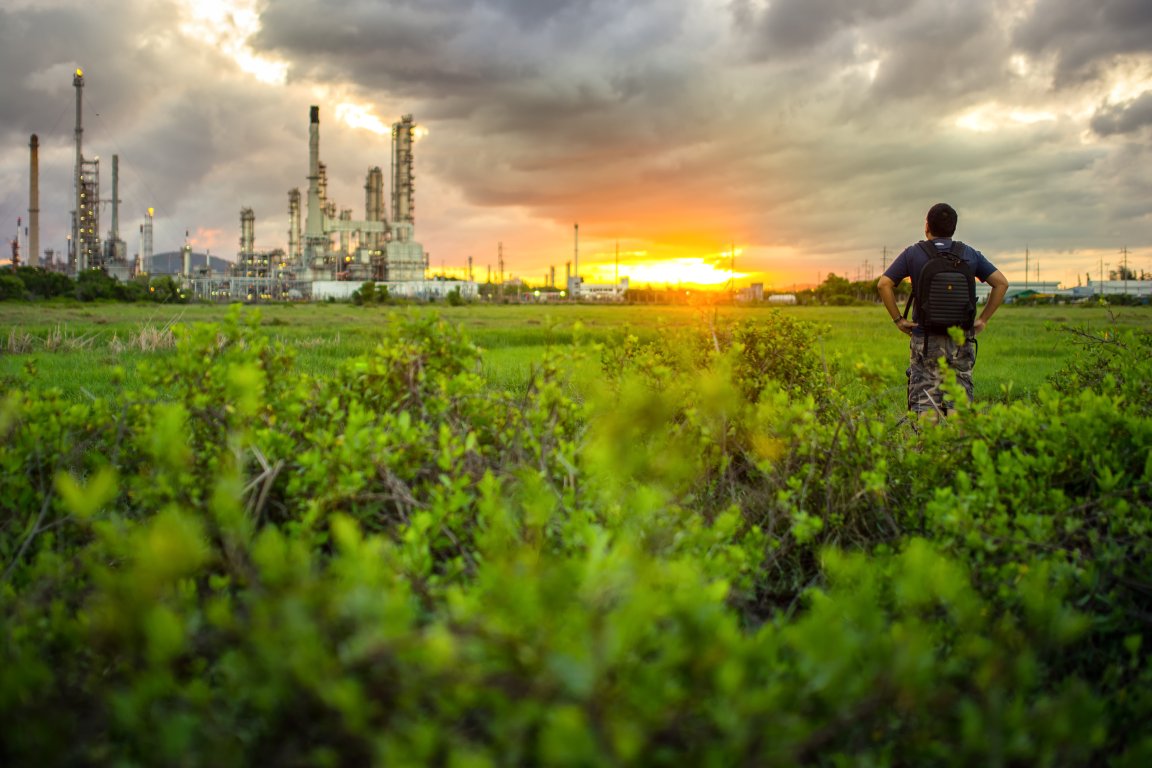 Man standing on a lush green field looking over an industrial refinery in the distance with a cloudy sky during sunset.