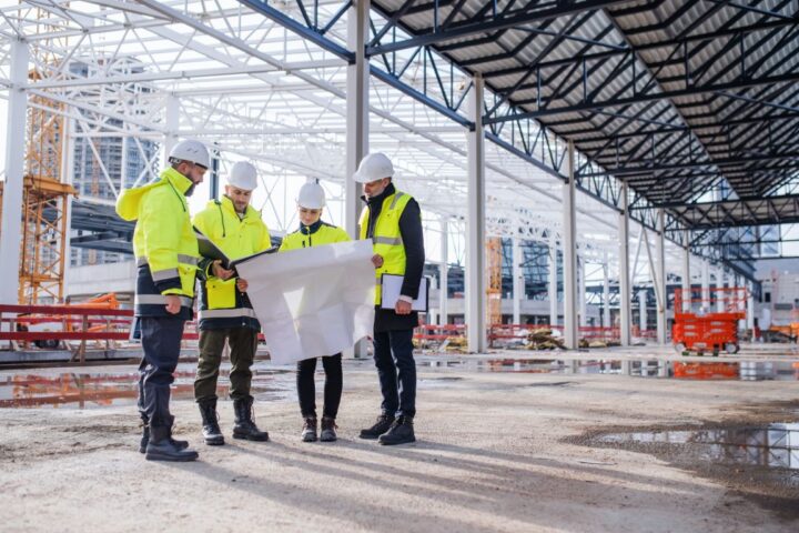 A group of four workers in high-visibility vests and jackets and white hard hats looking at blueprints together.