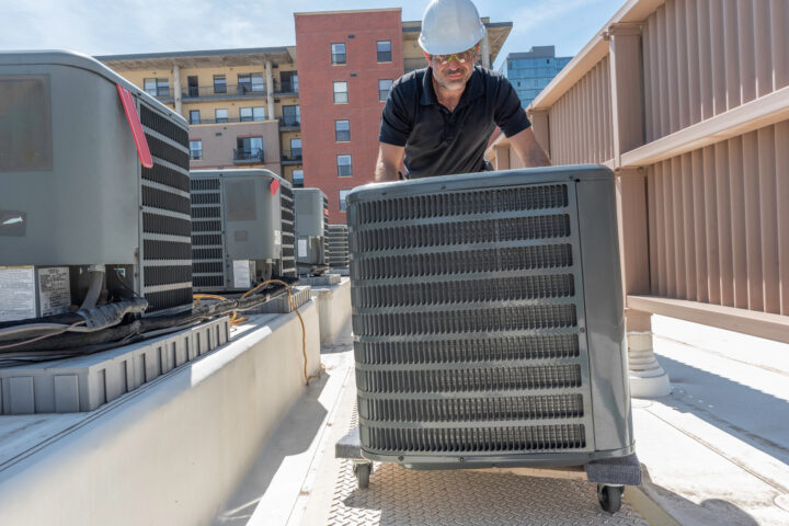 A worker rolls an HVAC unit on a pallet with wheels towards other air conditioners with tall buildings in the background.